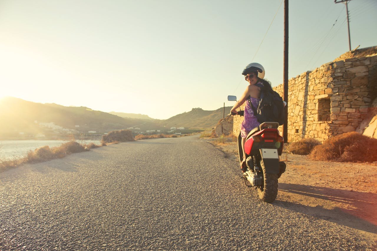 Woman riding scooter on scenic Greek road at sunrise, capturing adventure and travel vibes.